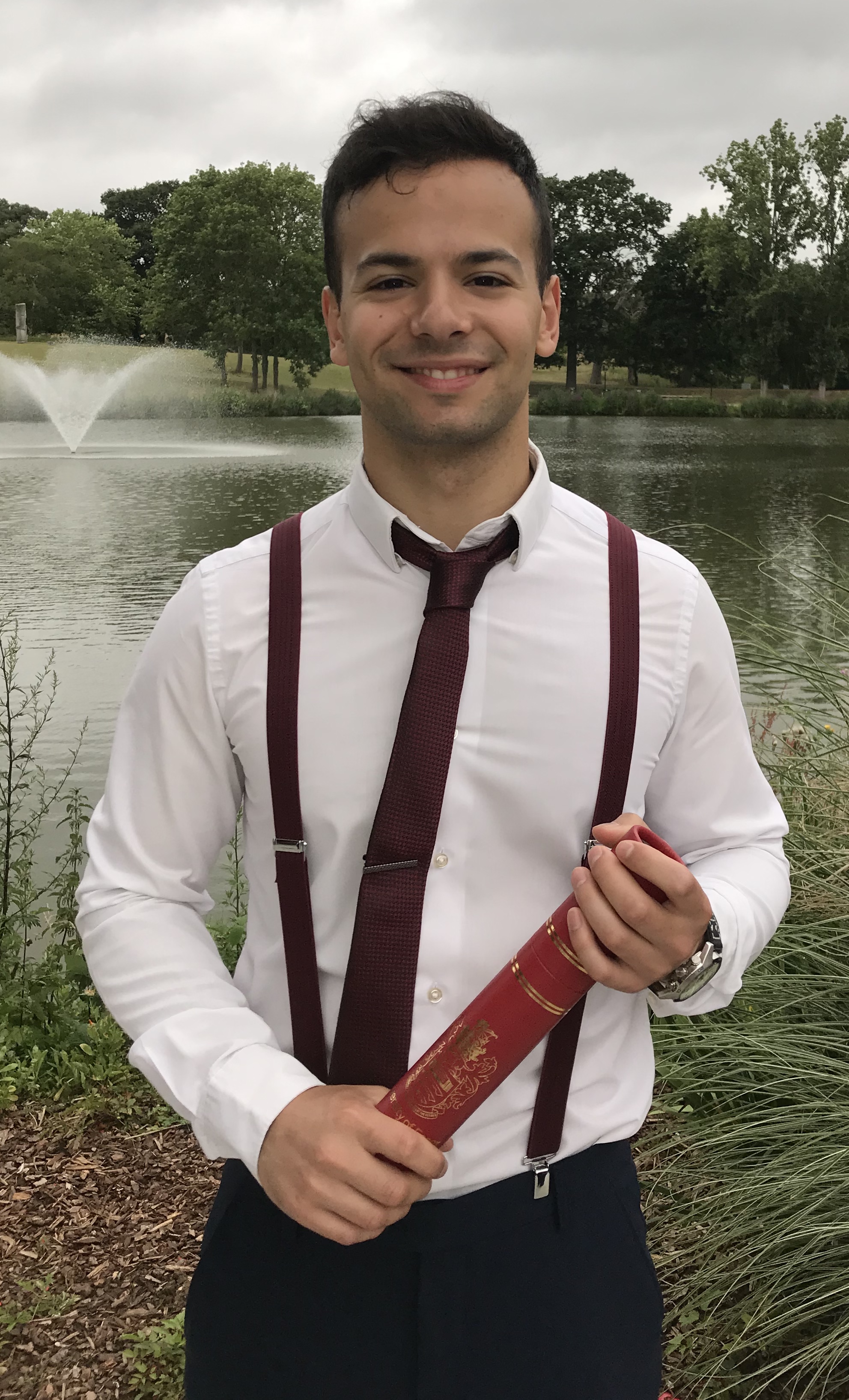 A picture of Giovanni Grasso standing in front of a lake in a white shirt with red tie & braces, holding his graduation certificate